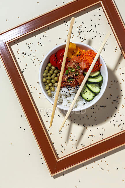 Von oben weiße Schüssel mit leckeren Poke-Gericht und Essstäbchen hinter Rahmen auf Tisch mit Sesam bedeckt platziert — Stockfoto