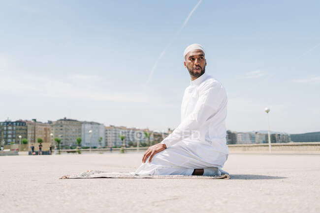 Full boy Islamic male in traditional white clothes standing on rug and praying against blue sky — Stock Photo