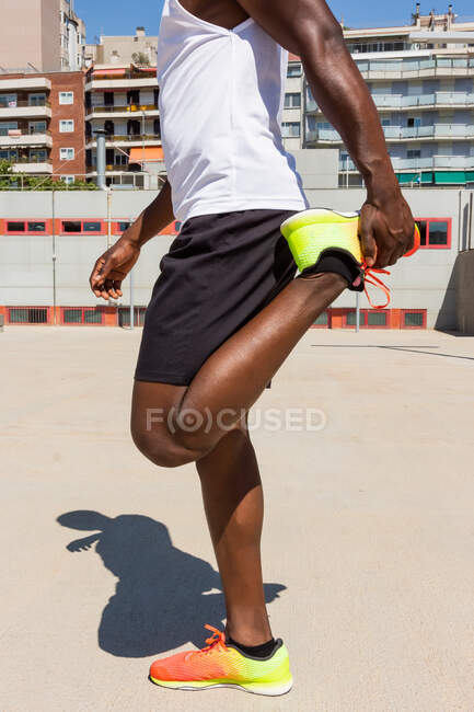 Vista lateral del atleta negro en forma que estira las piernas mientras calienta los músculos antes del entrenamiento en el campo de deportes en el día soleado - foto de stock