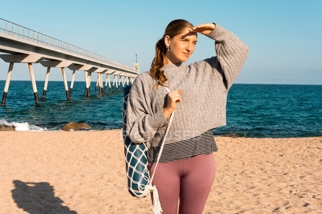 Joven hembra descalza en ropa deportiva con estera de yoga enrollada mirando a la distancia mientras está parada en la playa de arena cerca del mar - foto de stock