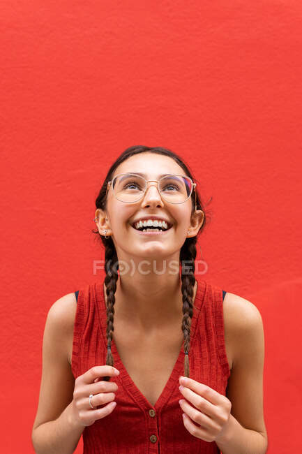 Cheerful young female touching pigtails while looking up on red background in street — Stock Photo