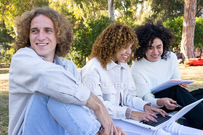 Hombre y mujeres multiétnicos con el pelo rizado sentado en el césped en el parque usando el ordenador portátil y compartiendo bloc de notas - foto de stock