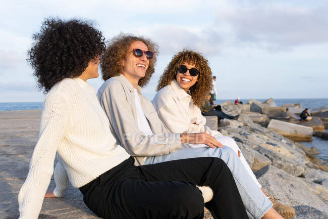 Side view of cheerful friends with curly hair wearing sunglasses sitting close against cityscape and rocky embankment in sunlight — Stock Photo