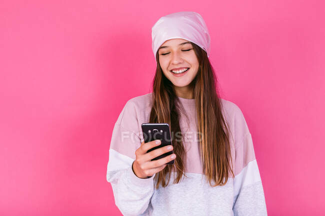 Adolescente feliz do sexo feminino com cabelo castanho no lenço de cabeça para a consciência conceito de câncer navegando no celular — Fotografia de Stock