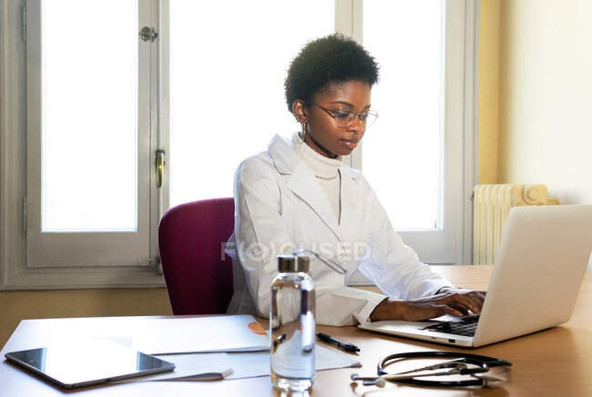 Молода афроамериканська жінка - лікар, яка друкує звіт про ноутбук, працюючи за столом у сучасному клінічному бюро. — стокове фото