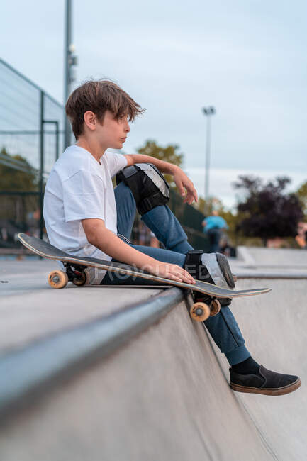 Вид сбоку мальчика-подростка, сидящего со скейтбордом на рейнджерах в скейт-парке и отводящего взгляд — стоковое фото