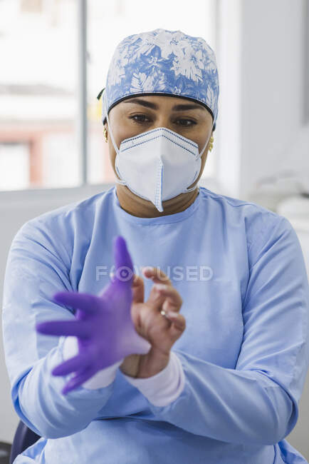Médico femenino en máscara respiratoria que se pone un guante de látex mientras mira hacia otro lado en el trabajo en el hospital - foto de stock
