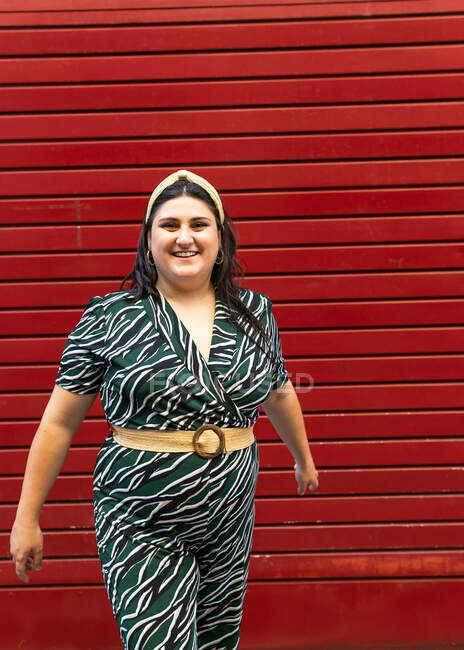 Портрет веселой юной брюнетки в стильной полосатой одежде и повязке при взгляде на камеру у красной стены — стоковое фото