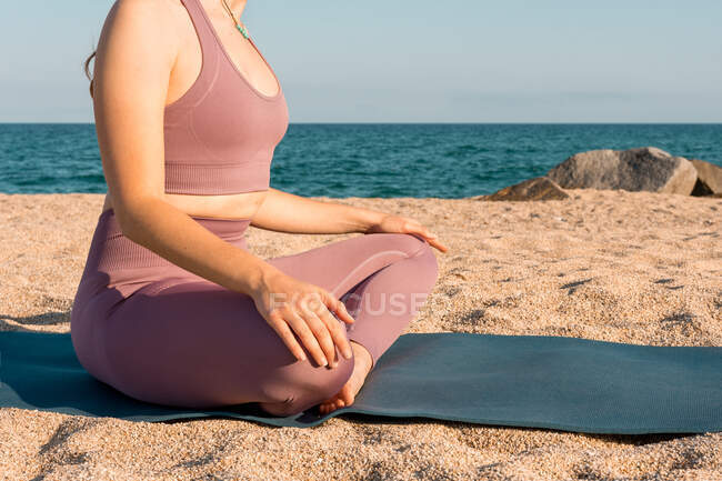 Vista lateral de una hembra serena irreconocible recortada sentada en Padmasana y haciendo yoga mientras medita con los ojos cerrados en la orilla del mar arenosa - foto de stock