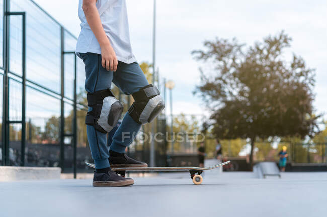Crop Niño adolescente en equipo de protección de pie con monopatín en el parque de skate y mirando hacia otro lado - foto de stock