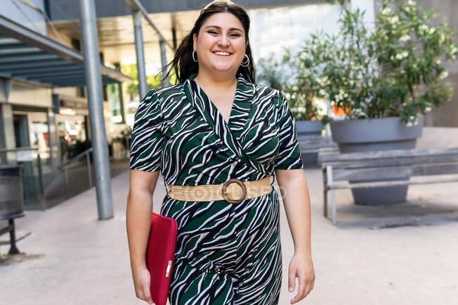 Moderne junge Plus-Size-Business-Lady in stylischem Kleid mit geometrischem Print und Gürtel, die Laptoptasche trägt, während sie auf urbanem Hintergrund läuft — Stockfoto