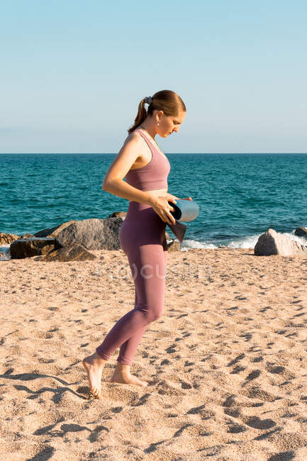 Полный вид сбоку тела молодой женщины в спортивной одежде кладет коврик для йоги на песок во время подготовки к тренировке на пляже возле океана — стоковое фото