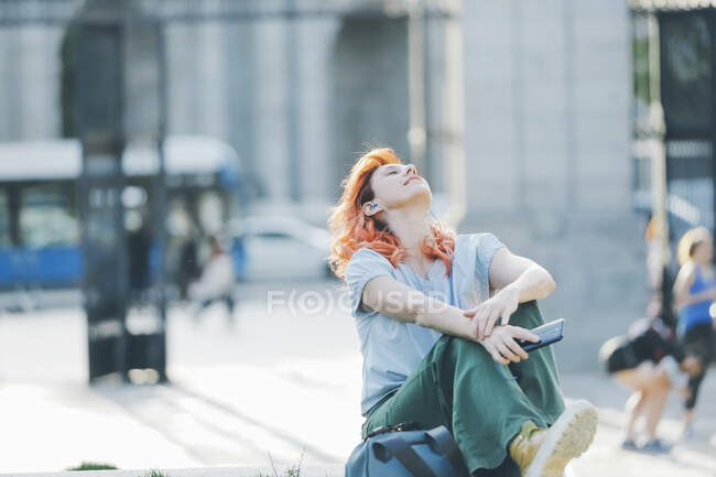 Femme rêveuse avec les cheveux roux assis dans la rue et écoutant de la musique dans les écouteurs tout en appréciant les chansons avec les yeux fermés et les bras tendus — Photo de stock