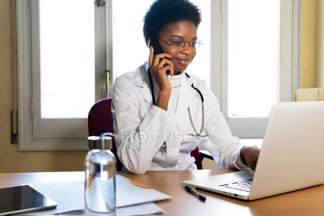 Competent young African American female medical practitioner answering phone call and using laptop while consulting patients remotely from office — Stock Photo