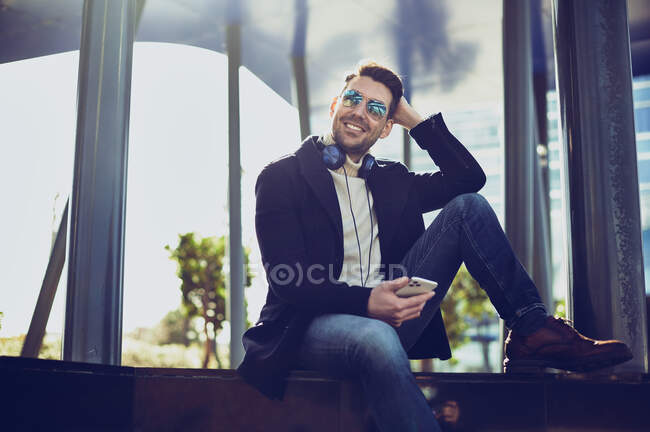Contenuto maschile in abbigliamento elegante con cuffie e cellulare seduto in città nella giornata di sole — Foto stock