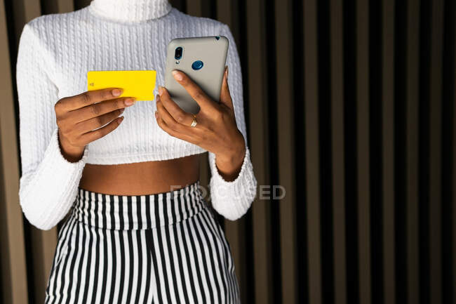 Нерозбірлива молода афроамериканська жінка в модному одязі, використовуючи кредитну картку і смартфон, щоб заплатити за онлайн-замовлення проти смугастих стін на вулиці — стокове фото