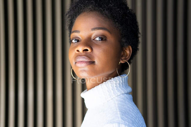 Seitenansicht einer emotionslosen Afroamerikanerin im trendigen Pullover, die in die Kamera gegen eine gestreifte Hauswand auf der Straße blickt — Stockfoto