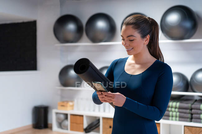 Mujer joven positiva en ropa deportiva con mensajería de estera de yoga enrollada en el teléfono móvil después de la práctica en un moderno estudio de fitness - foto de stock
