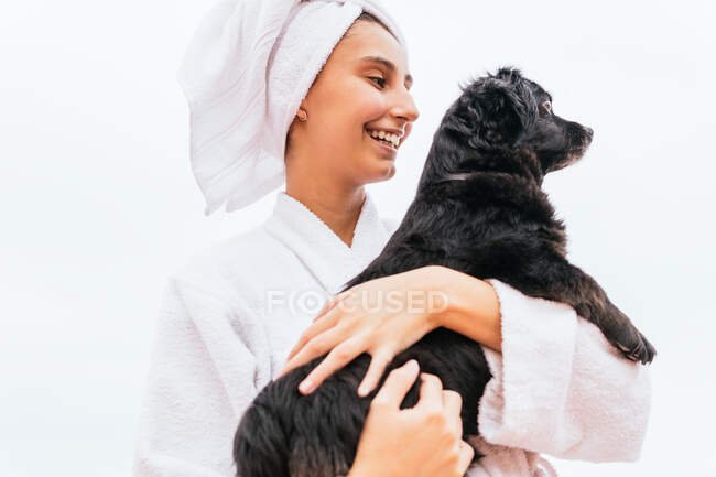 Jovem otimista em roupão de banho e toalha sorrindo e abraçando o cachorro preto durante a sessão de spa em casa — Fotografia de Stock