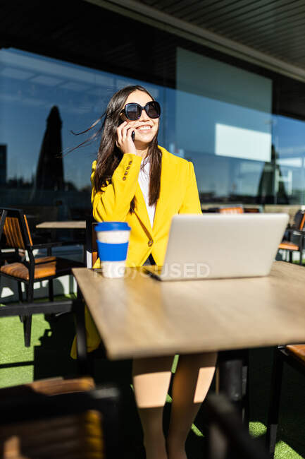 Sorridente donna d'affari asiatica con cappotto giallo seduta a un tavolo a prendere il caffè con il suo smartphone e laptop — Foto stock