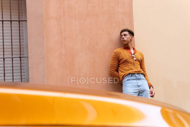 Joven chico de pelo rizado étnico con estilo en traje de moda apoyado contra la pared cerca de aparcado moderno automóvil naranja en la calle urbana - foto de stock