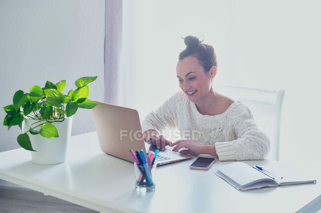 Весела жінка віддаленого працівника, що серфінгує Інтернетом на нетбуці за столом з смартфоном і копіювальною книгою вдома на сонячному світлі — стокове фото