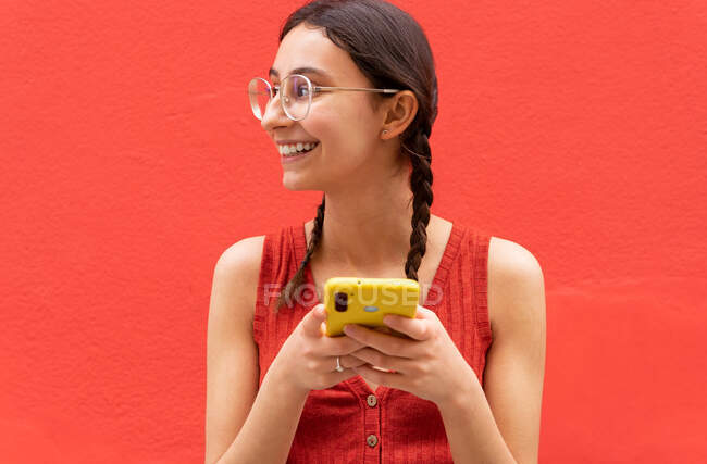 Jovem alegre em pigtails penteado navegando no smartphone de pé olhando para longe no fundo vermelho na rua — Fotografia de Stock