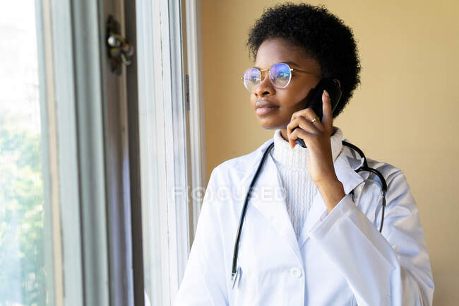 Young African American female doctor in white coat with stethoscope talking mobile phone while standing near window in clinic — Stock Photo