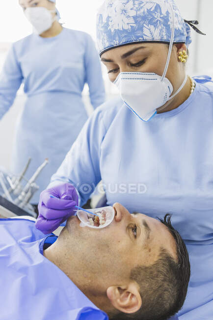 Spécialiste dentaire ethnique professionnel portant l'uniforme médical faisant la procédure de durcissement au client en clinique avec infirmière — Photo de stock