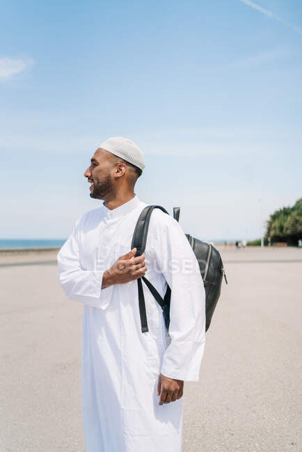Homem islâmico feliz em roupas tradicionais ajustando mochila e olhando para longe com sorriso enquanto passa o dia ensolarado de verão na praia — Fotografia de Stock