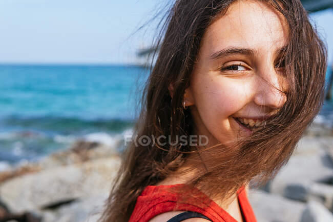 Позитивна жінка в стильному вбранні стоїть на камені на березі моря і насолоджується сонячним днем під час літніх канікул дивлячись на камеру — стокове фото