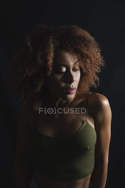 Charming African American female model with curly hair looking down in dark studio — Stock Photo