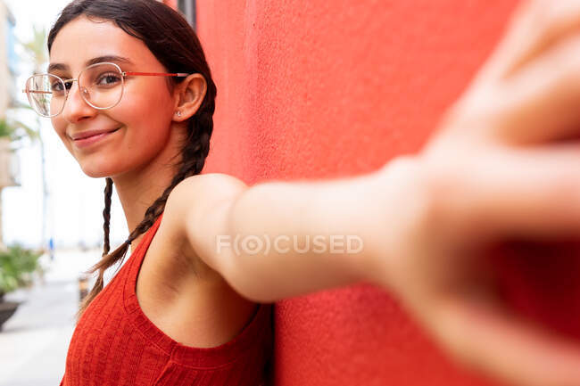 Side view of content female in trendy glasses leaning on red wall of building in street and looking at camera with outstretched arm — Stock Photo