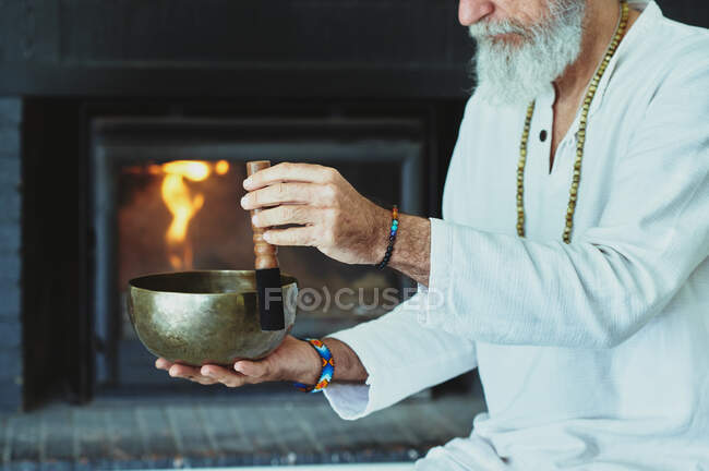 Varón anciano con barba gris jugando a cantar con un delantero de madera mientras mira hacia otro lado durante la práctica espiritual - foto de stock