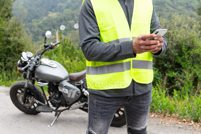 Crop ciclista masculino en chaleco de mensajería en el teléfono móvil, mientras que de pie cerca de la motocicleta rota cerca de exuberantes bosques verdes - foto de stock