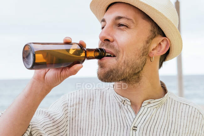 Счастливый молодой бородатый хипстер мужчина путешественник в стильной летней рубашке и шляпе наслаждаясь холодным пивом из бутылки во время отдыха на берегу моря во время летних каникул — стоковое фото