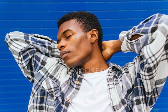 Homme afro-américain rêveur avec les mains derrière la tête et les yeux fermés debout sur fond de mur bleu dans la rue — Photo de stock