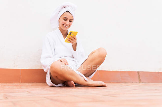 Молода жінка в халаті і рушнику посміхається і переглядає мобільний телефон, відпочиваючи на балконі після душу — стокове фото