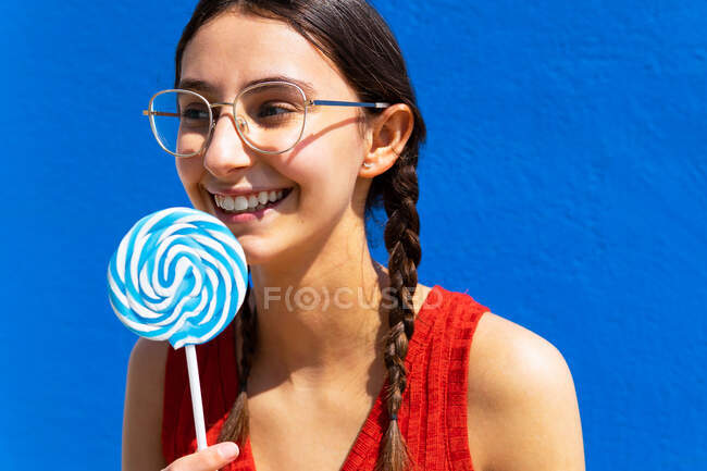 Positive charming female standing with sweet lollipop in street on sunny day on blue background and looking away — Stock Photo