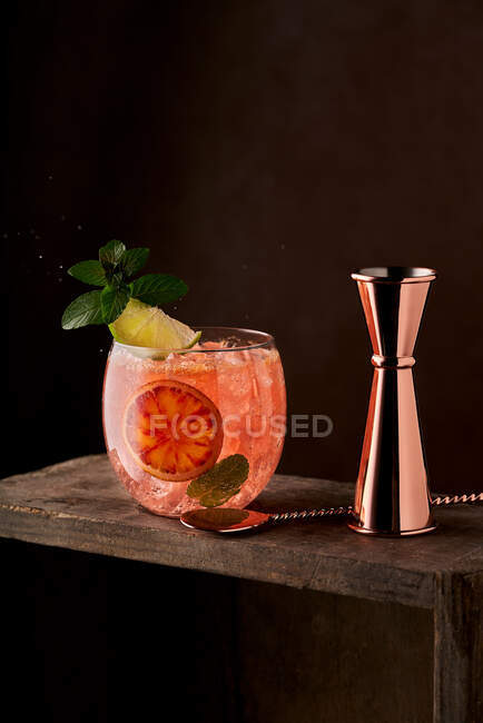Appetizing refreshing cocktail with ice cubes and citruses served on wooden table with metal jigger and spoon against black background — Stock Photo