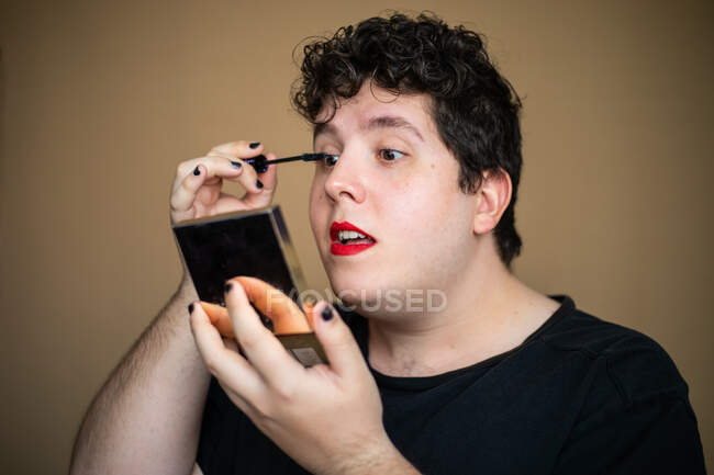 Концентрированный эксцентричный женственный мужчина наносит тушь с кистью, делая макияж с открытым ртом и держа зеркало — стоковое фото