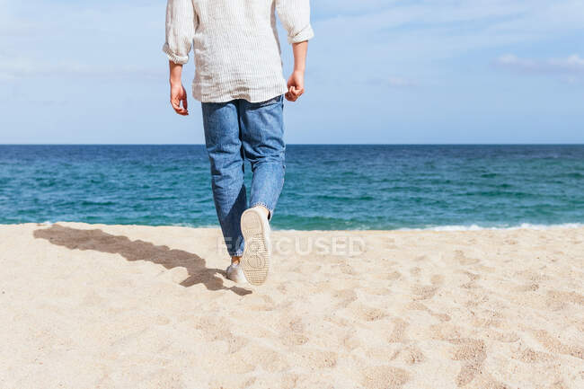 Вид ззаду на обрізаного невпізнаваного чоловіка в модному повсякденному одязі і кепці, що йде один на піщаному пляжі до махаючого моря, проводячи літні канікули на березі моря — стокове фото