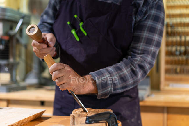 Unrecognizable male woodworker with wooden hammer and chisel carving wooden detail while working in professional joinery workshop — Stock Photo