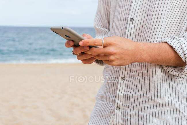 Cropped unrecognizable man in stylish shirt browsing mobile phone on sandy beach near sea — Stock Photo