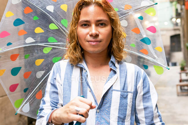 Zufriedener stylischer Mann mit langen Haaren, der an regnerischen Tagen unter einem durchsichtigen Regenschirm auf der Straße steht und in die Kamera blickt — Stockfoto