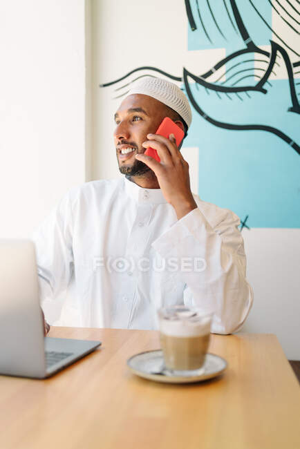 Hombre musulmán feliz en ropa auténtica sentado en la mesa y netbook de navegación en la cafetería - foto de stock