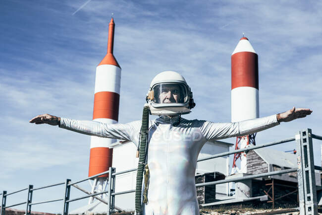 Man in spacesuit standing with outstretched arms on rocky ground against metal fence and striped rocket shaped antennas on sunny day — Stock Photo