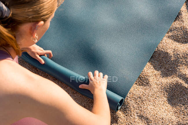 Сверху обрезанная неузнаваемая молодая женщина в спортивной одежде кладет коврик для йоги на песок во время подготовки к тренировке на пляже возле океана — стоковое фото