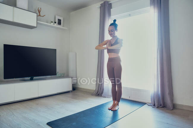 Молодая сконцентрированная самка в спортивной одежде с закрытыми глазами и руками намасте, стоящая на коврике для йоги дома в подсветке. — стоковое фото