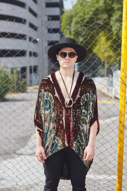 Впевнений самець в модних сонцезахисних окулярах і капелюсі стоїть біля сітки паркану на вулиці і дивиться на камеру — стокове фото